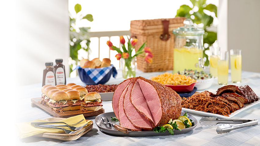 Honey Baked Bone-in Half Ham, Pulled Pork Cuban Sliders, BBQ Pork Roast and BBQ Pork Ribs with baked beans, double macaroni and cheese in serving dishes on a picnic table